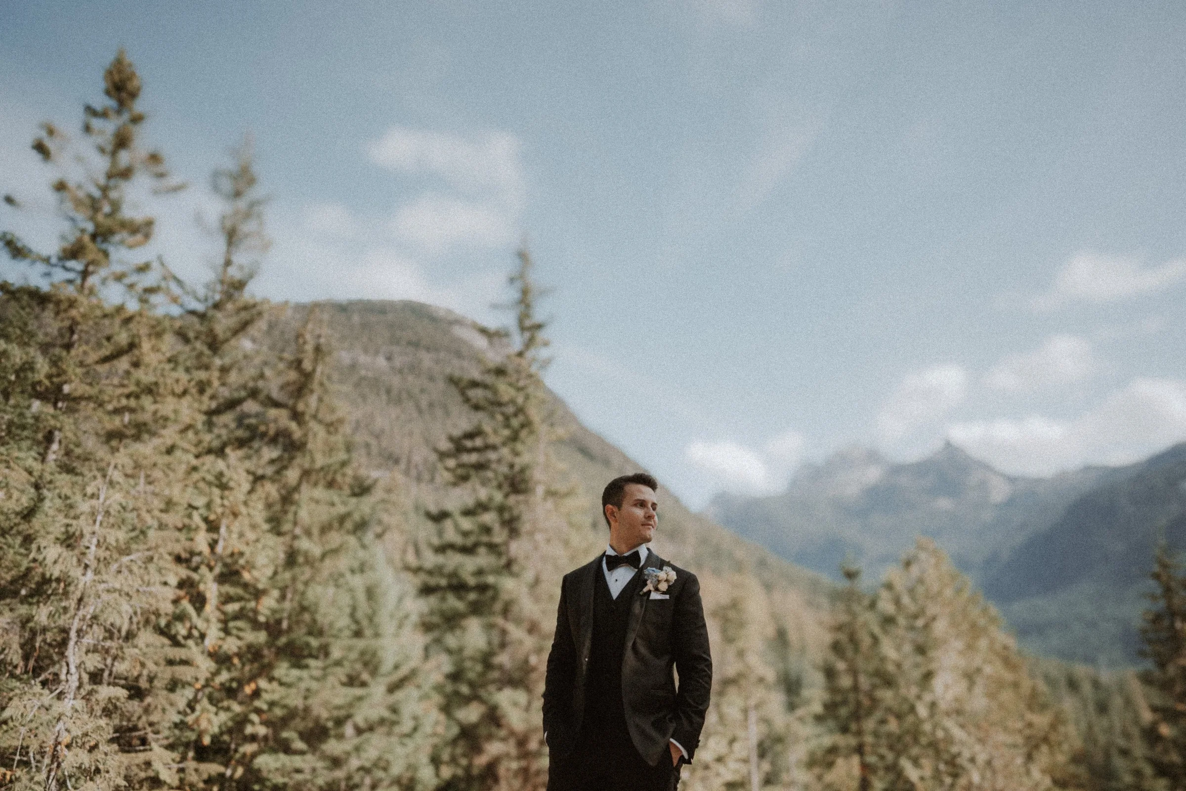 Groom standing alone with mountain peaks and forests behind him.