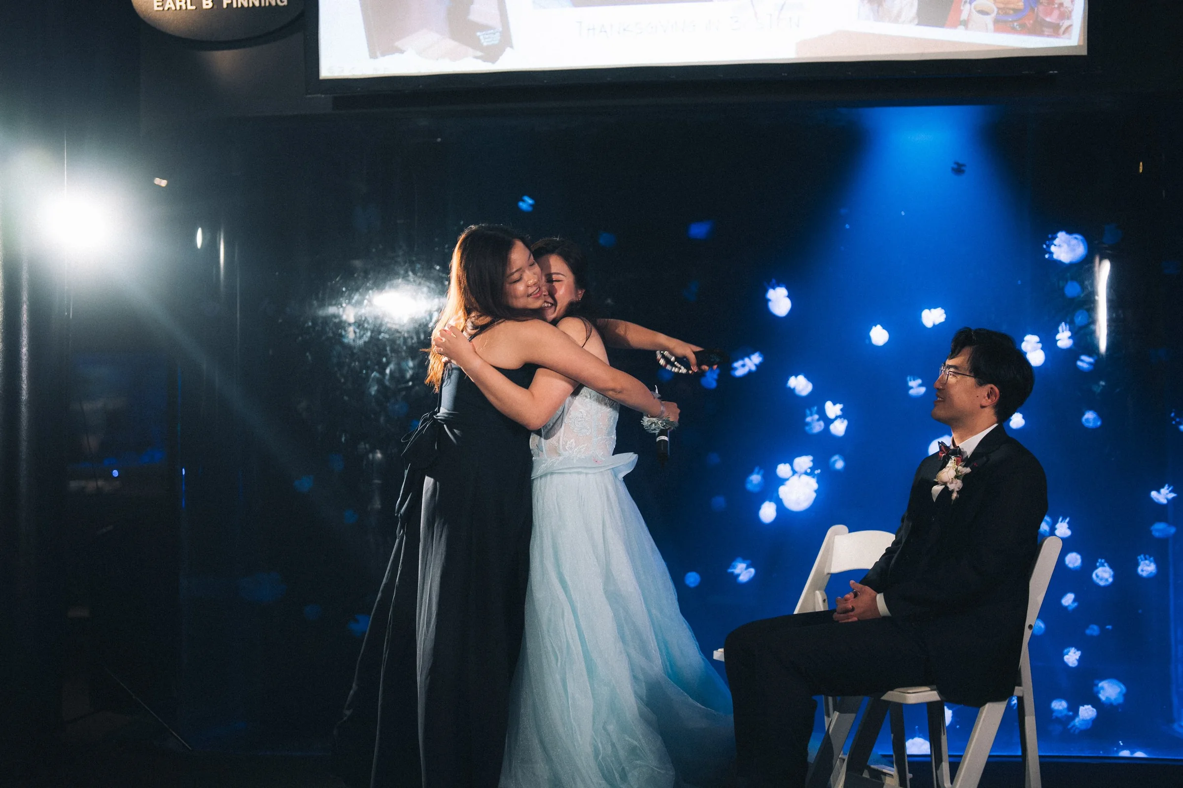 Bride hugging a guest while the groom watches during the reception.