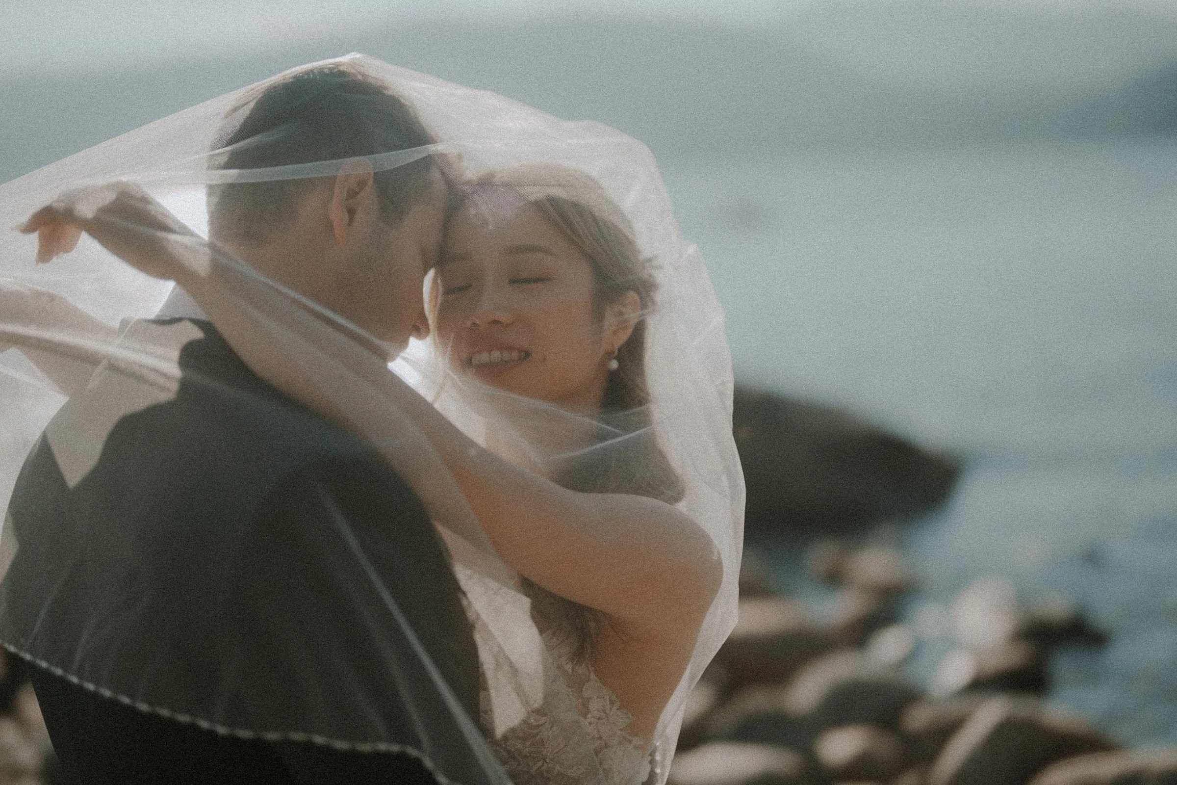 Couple kissing through a translucent veil beside the coast.