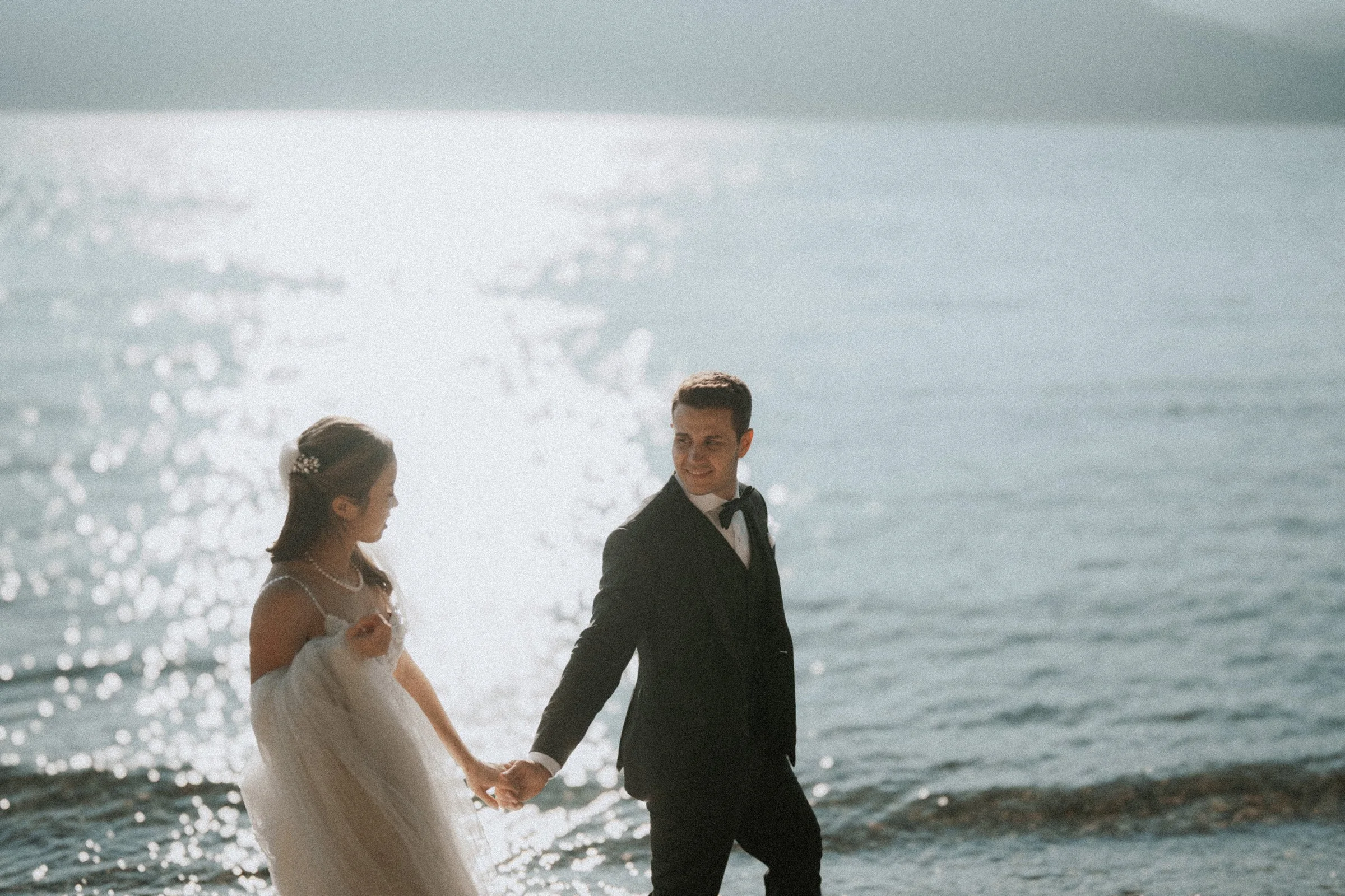 Bride and groom walking beside sparkling water on the pebbled beach.