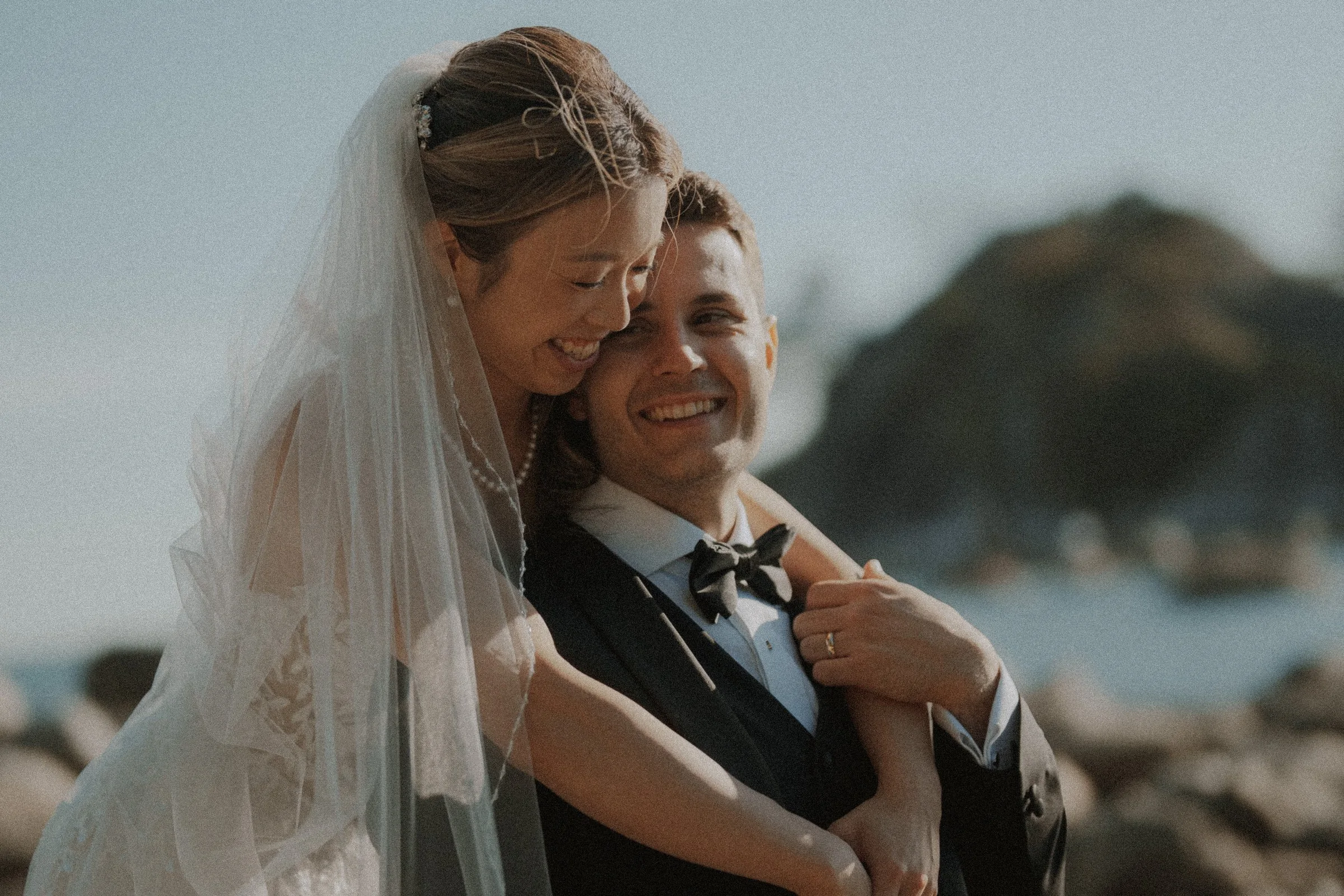 Bride embracing the groom from behind as they smile beside the coast.