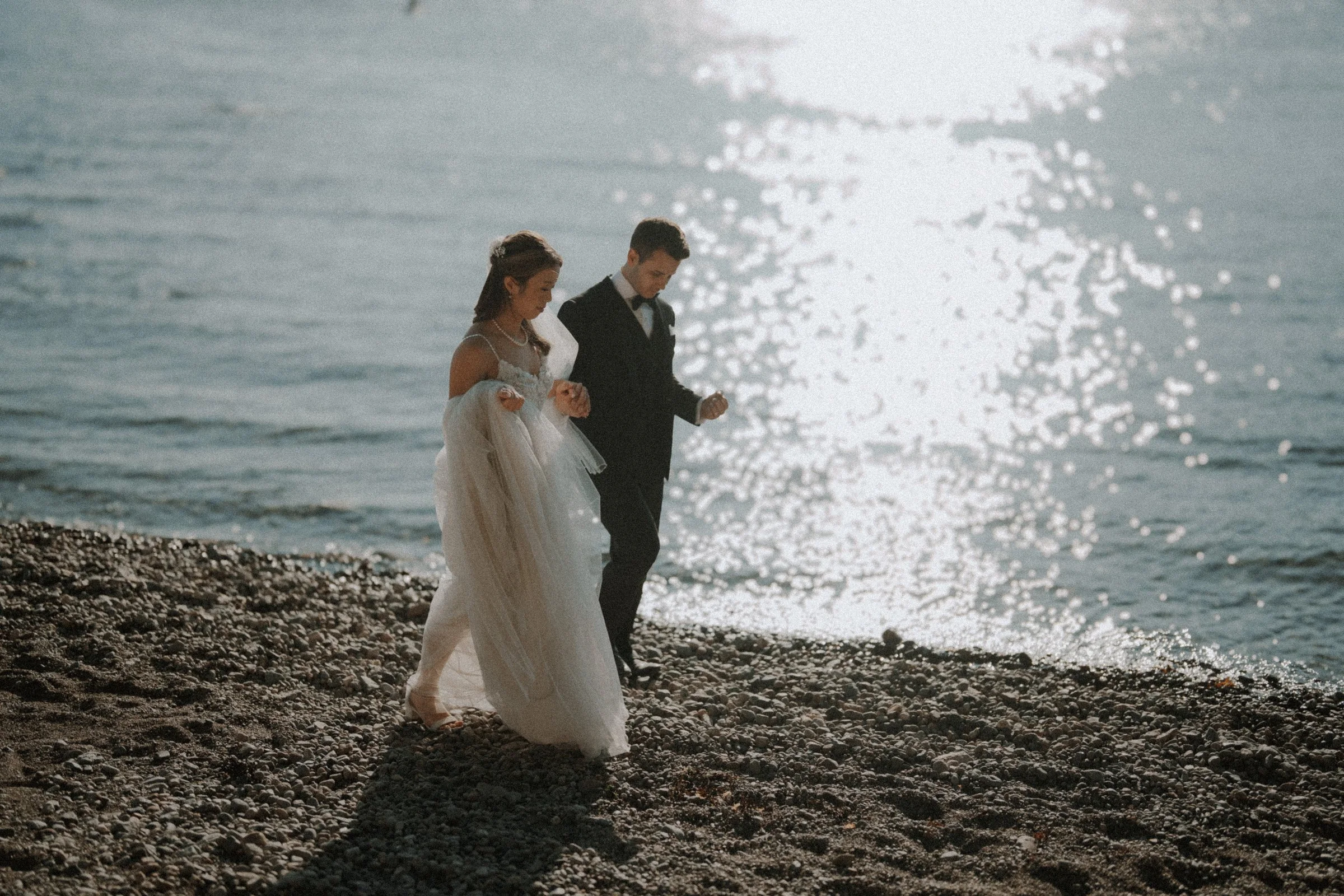 Bride and groom walking beside sparkling sunlit water.