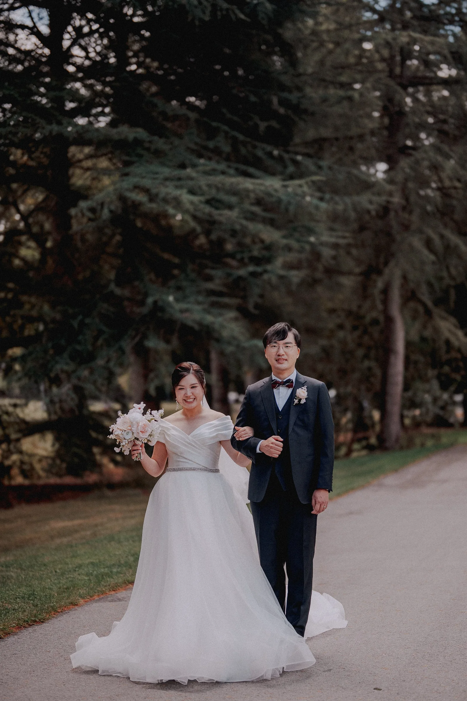 Bride and groom walking arm in arm along a tree-lined path.