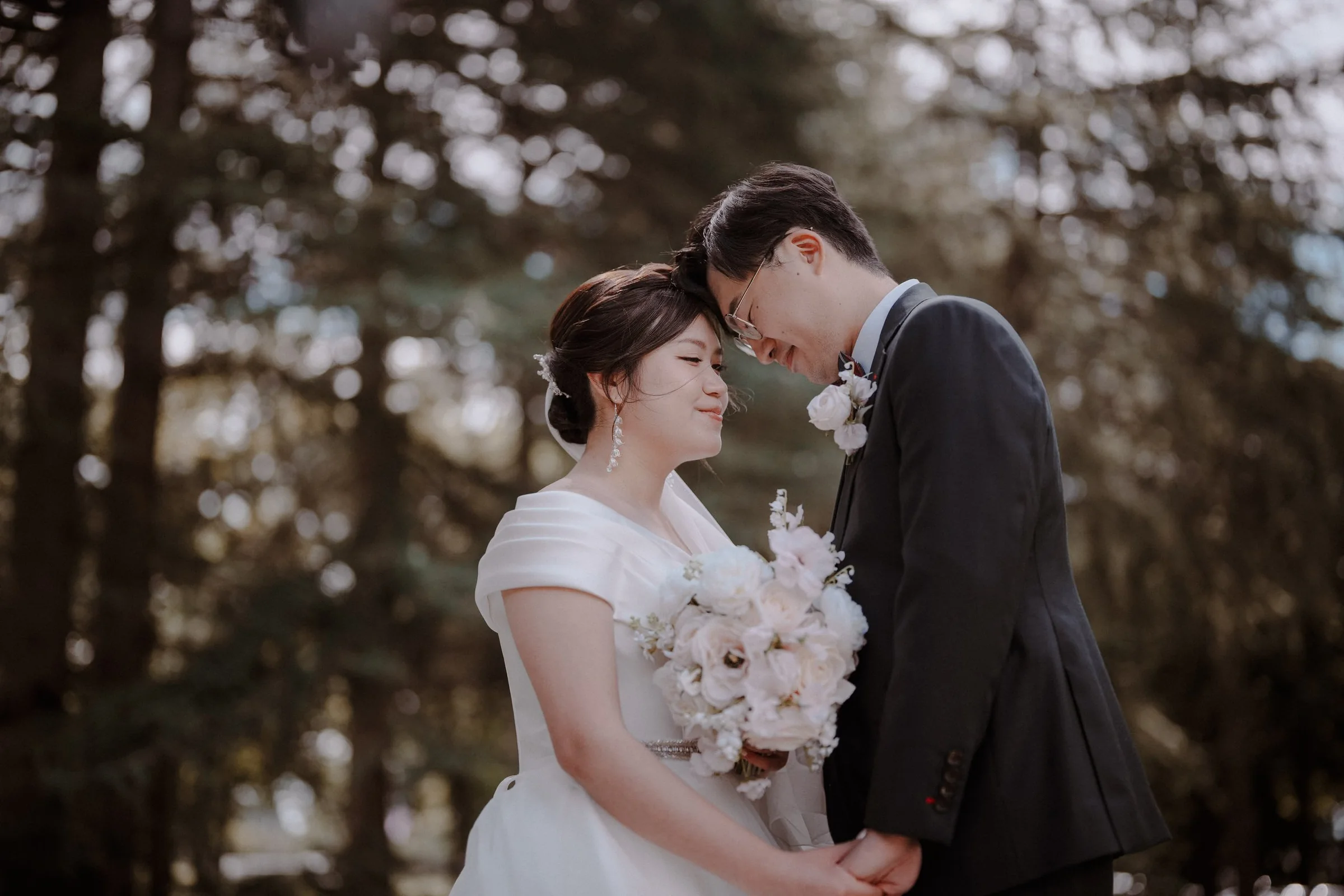 Couple touching foreheads beneath leafy trees after their wedding ceremony.