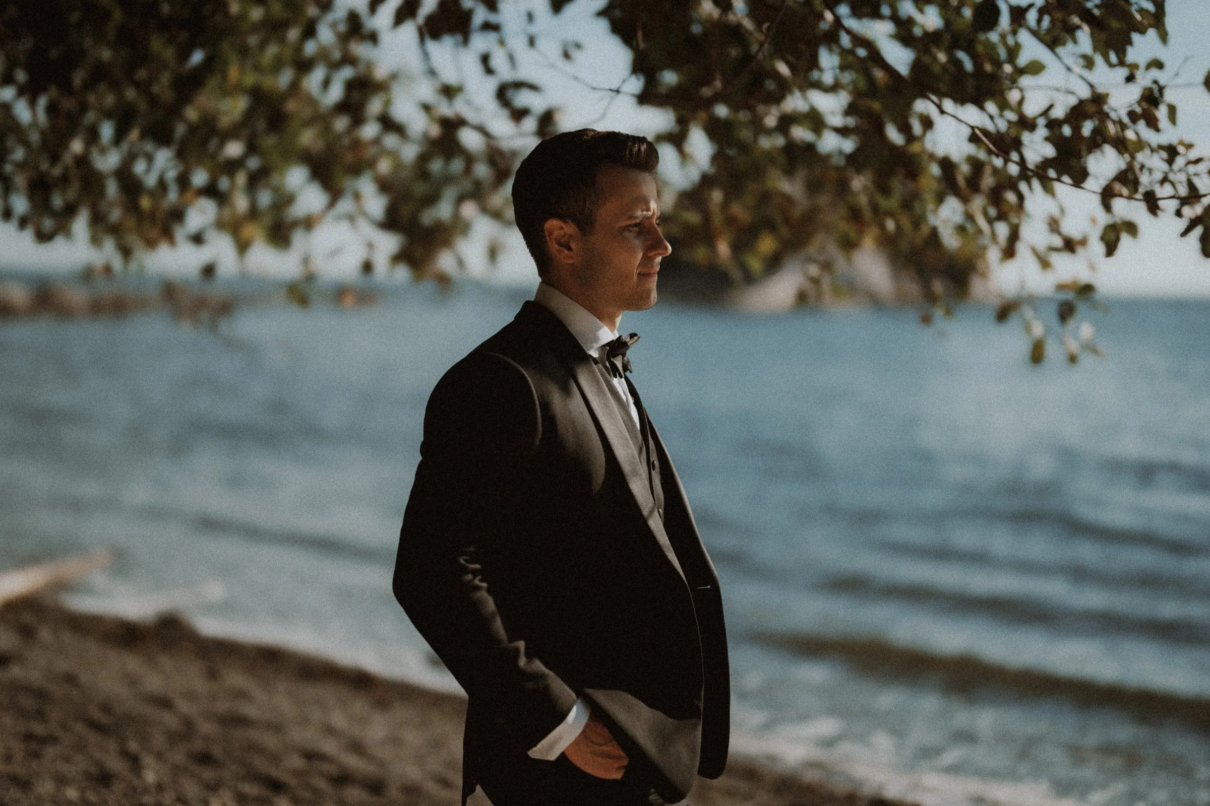 Portrait of the groom beneath leafy branches by the shoreline.