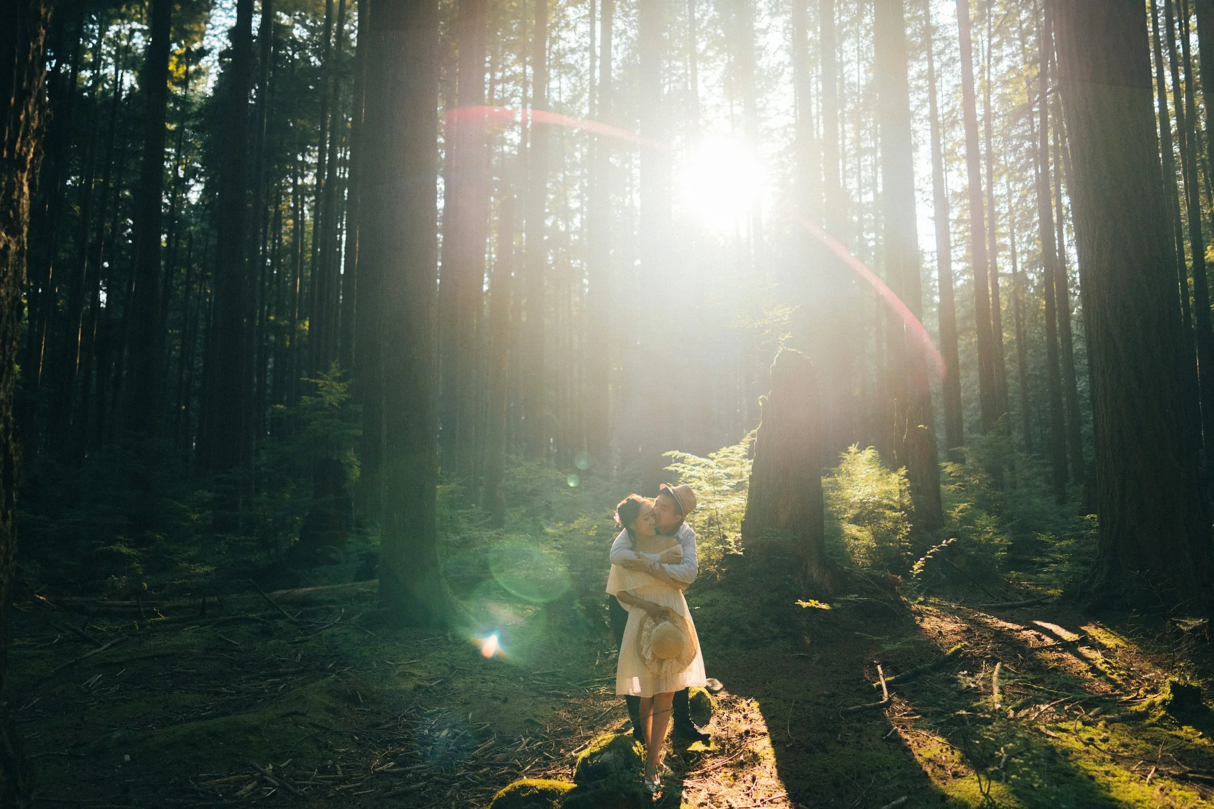 Candid couple hugging, strong sun flare, emotional forest engagement portrait.