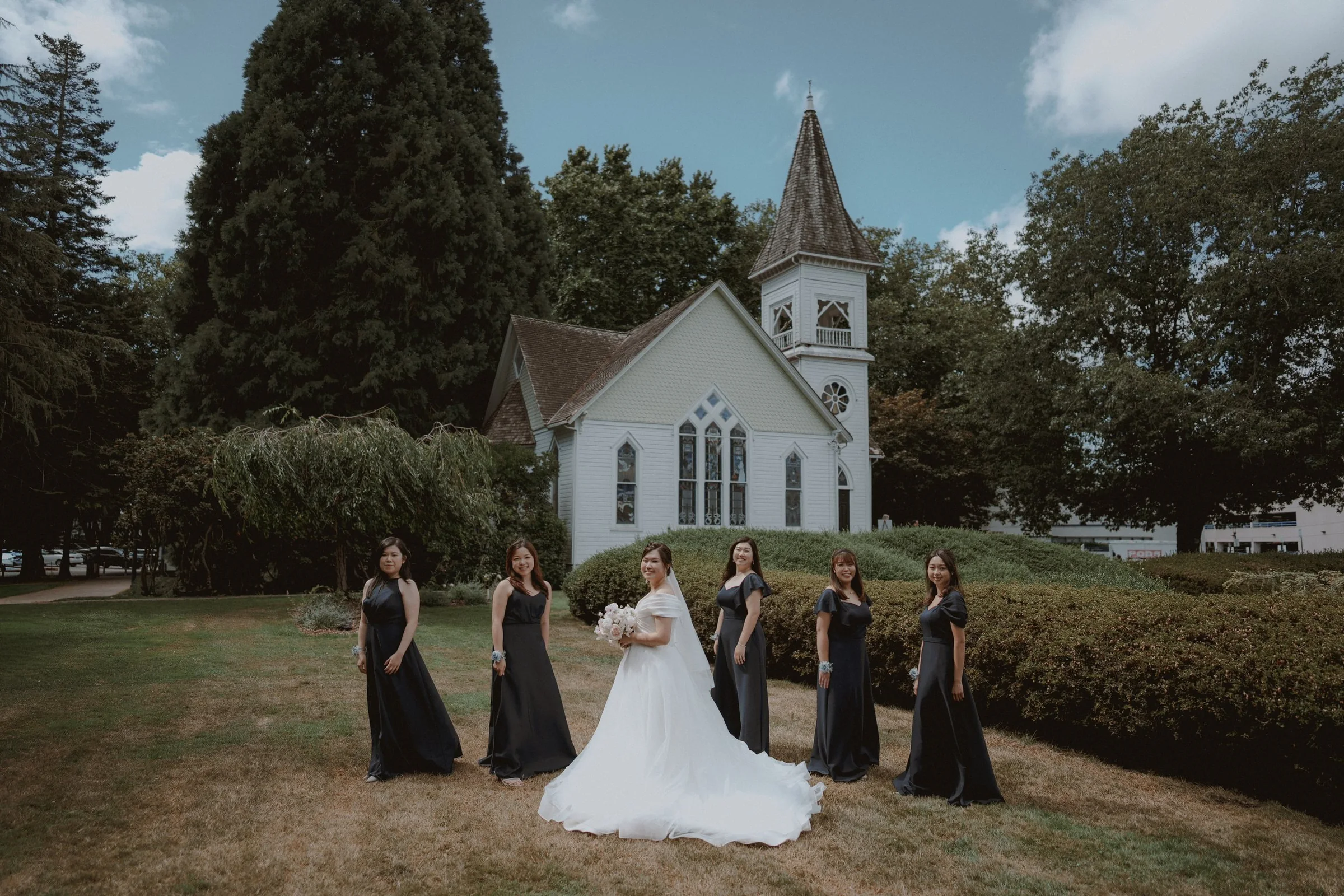 Bride with bridesmaids in black dresses on the Minoru Chapel lawn.