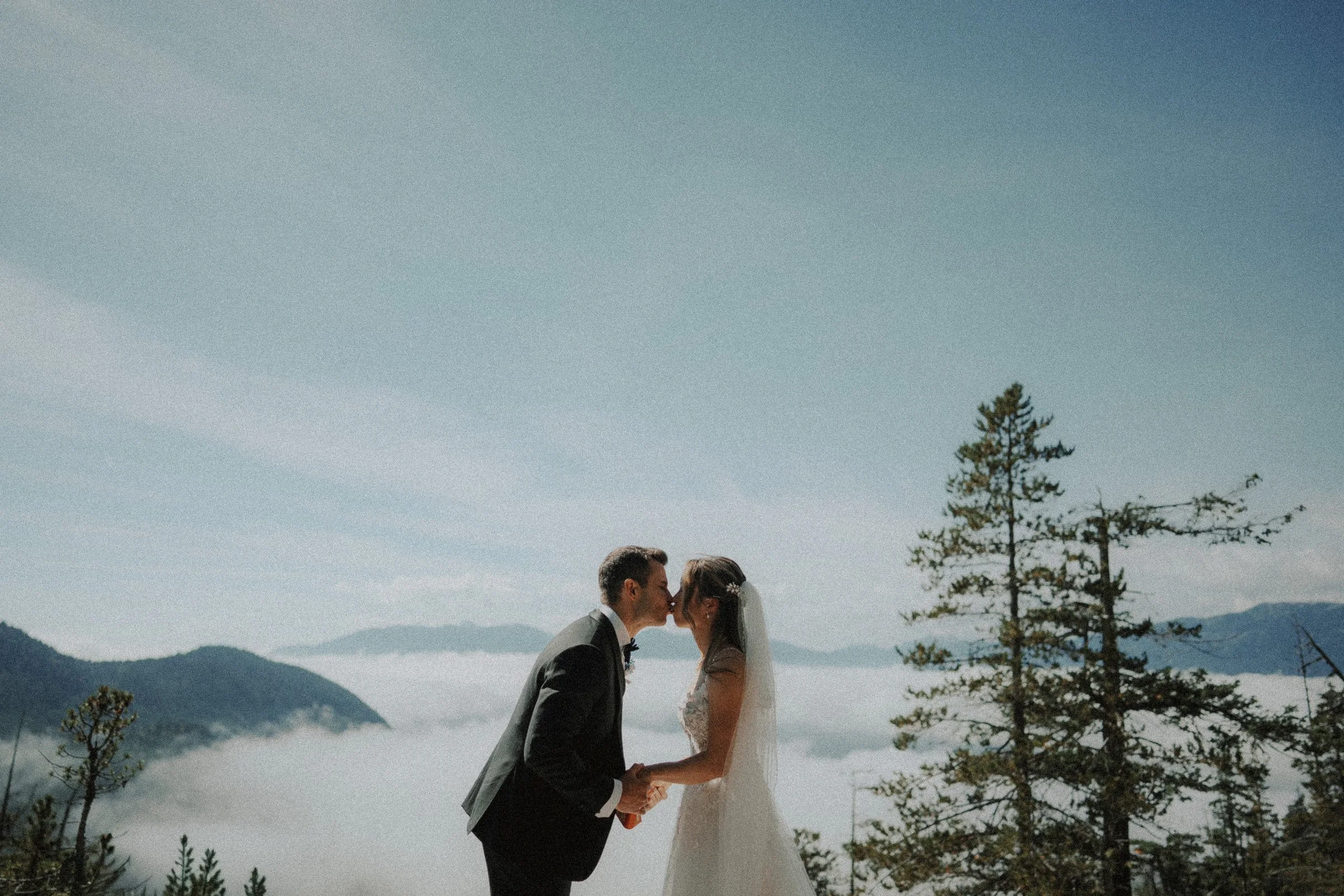 Bride and groom sharing a kiss above a cloud-filled mountain valley.
