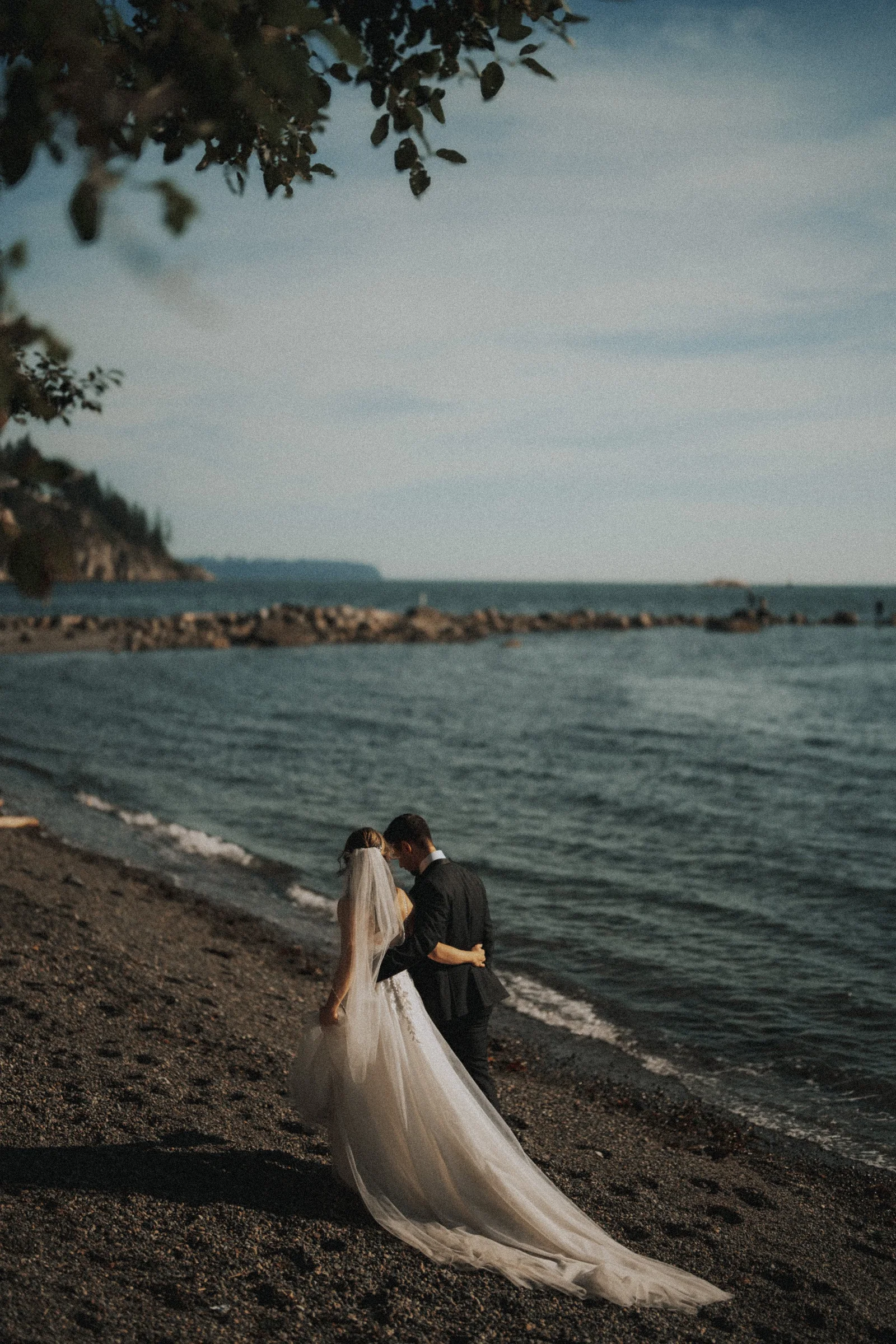 Bride and groom standing together beside the water beneath overhanging leaves.