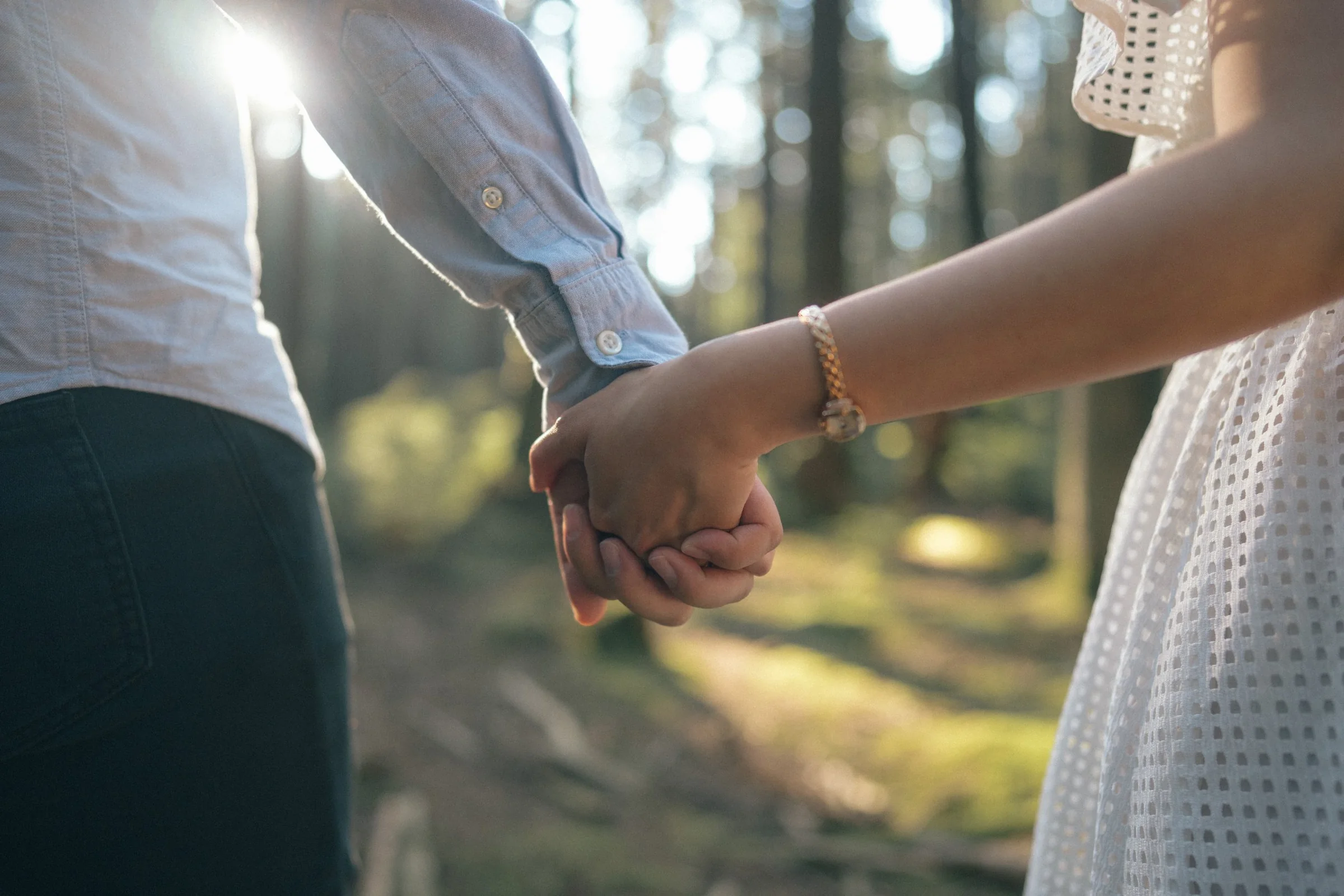 Close-up of couple holding hands, strong backlight, candid forest engagement detail.