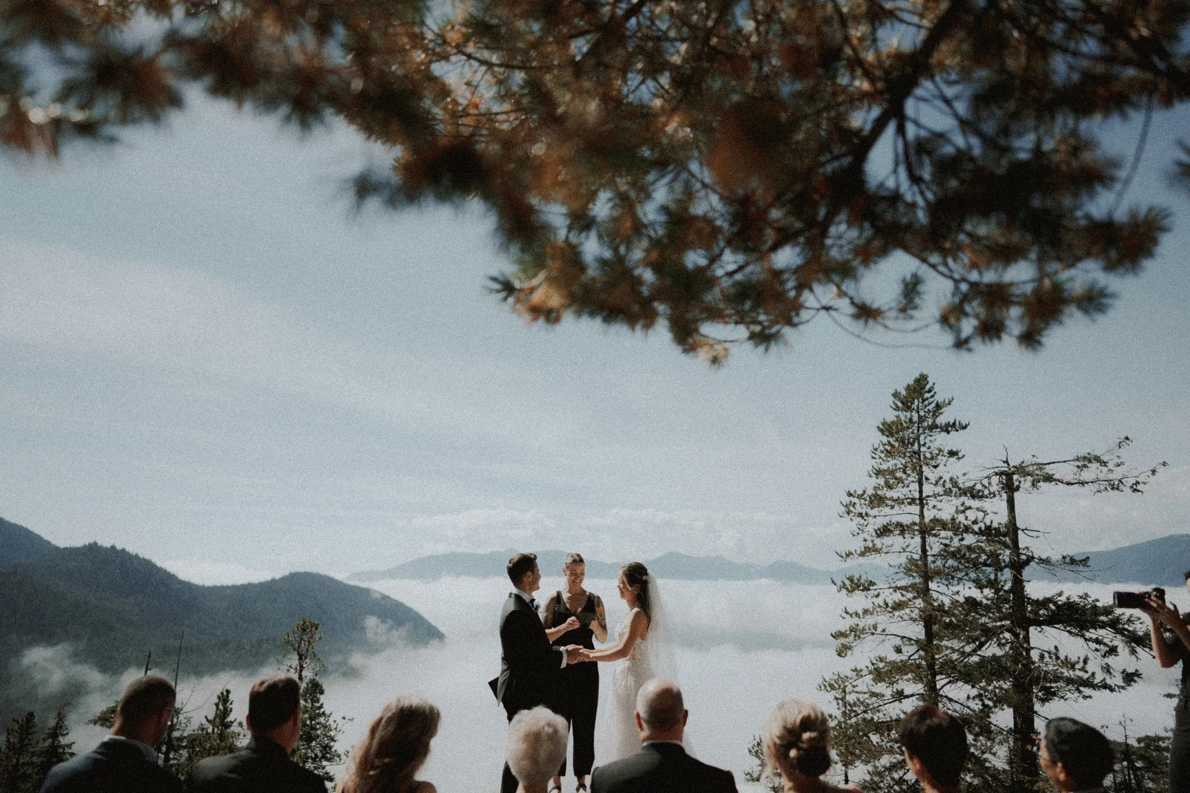 Couple holding hands during their vows above cloud-covered mountains.