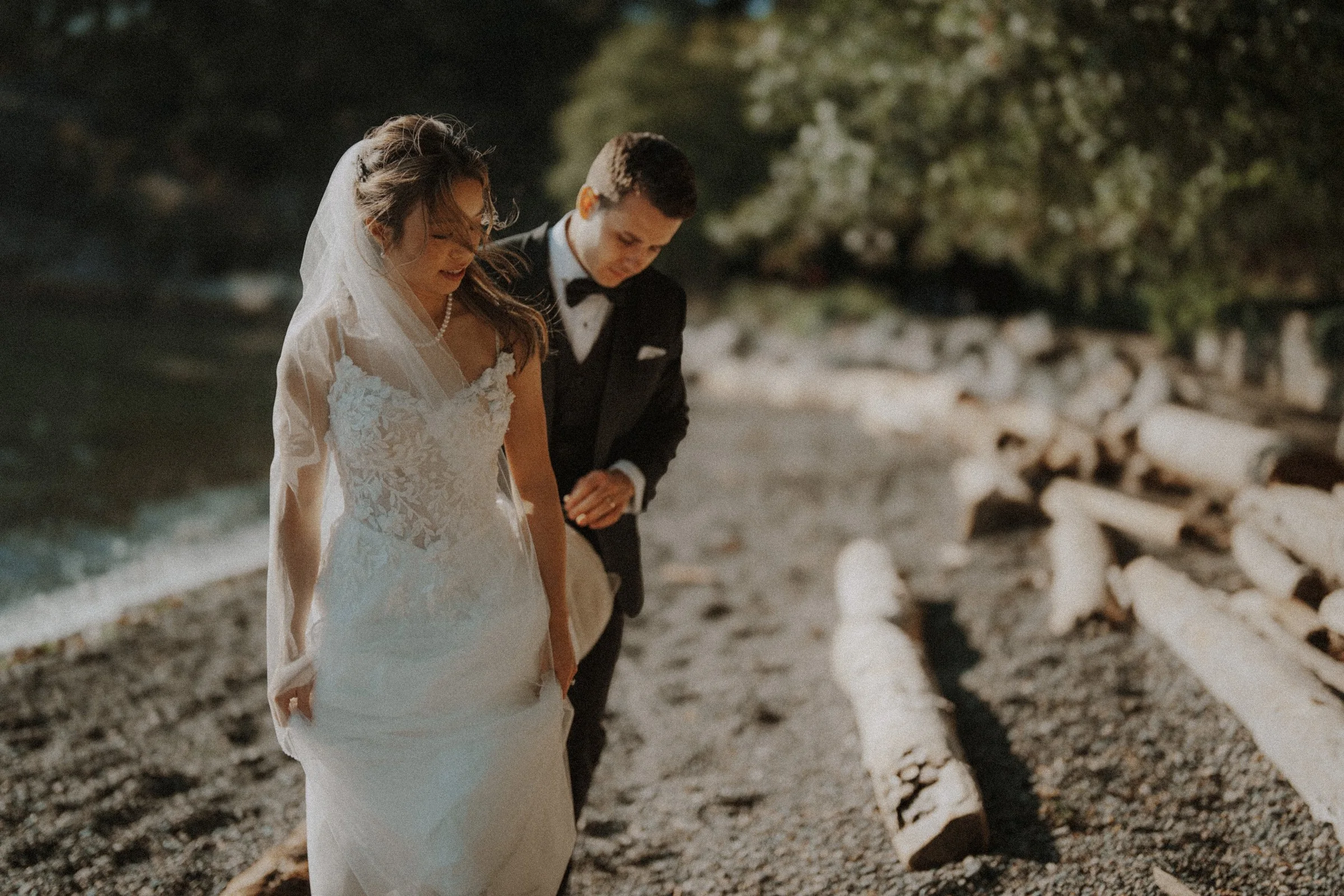 Couple walking together beside driftwood and sunlit water.