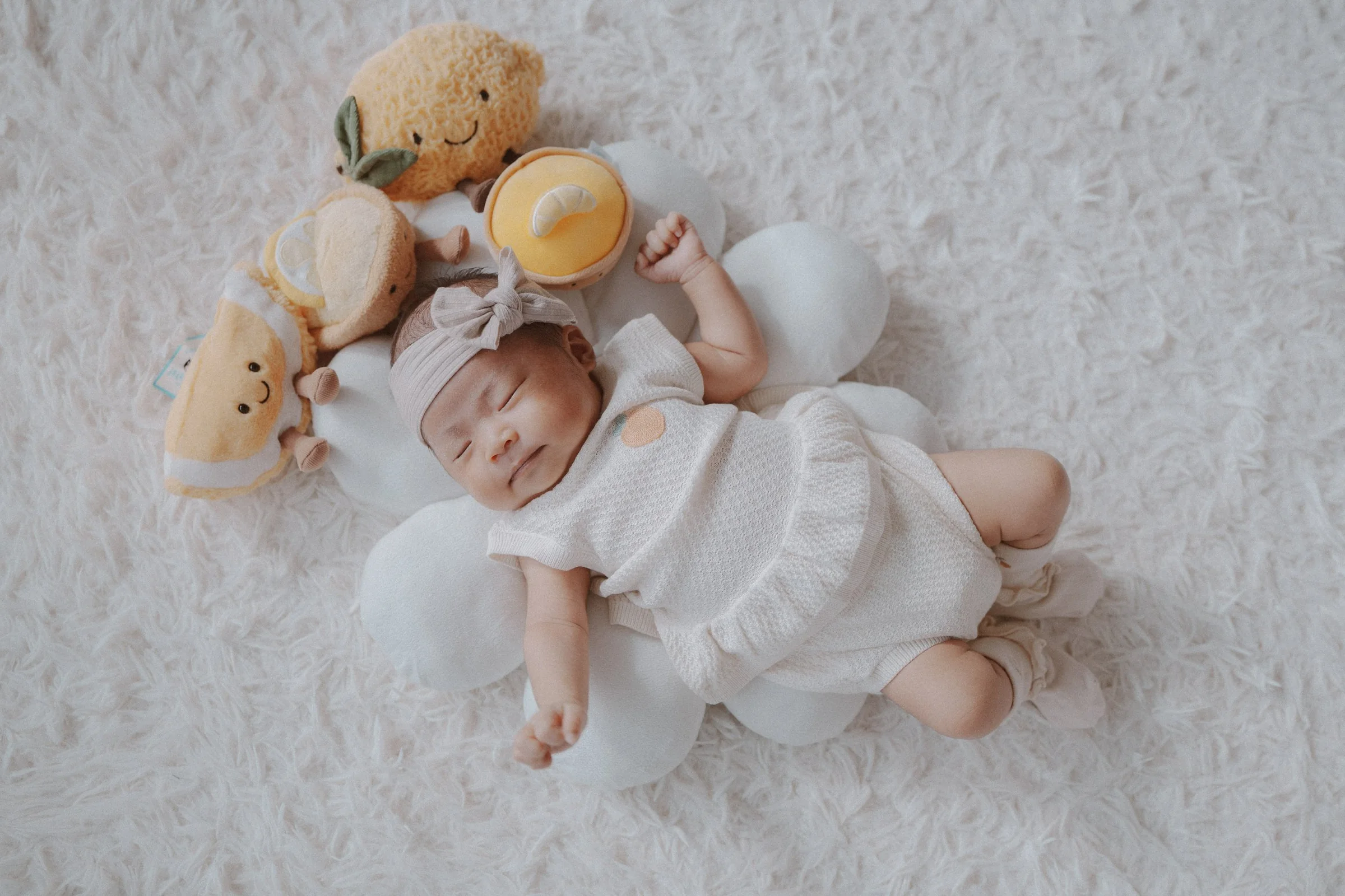 Sleeping newborn stretched out beside soft toys on a pale blanket.