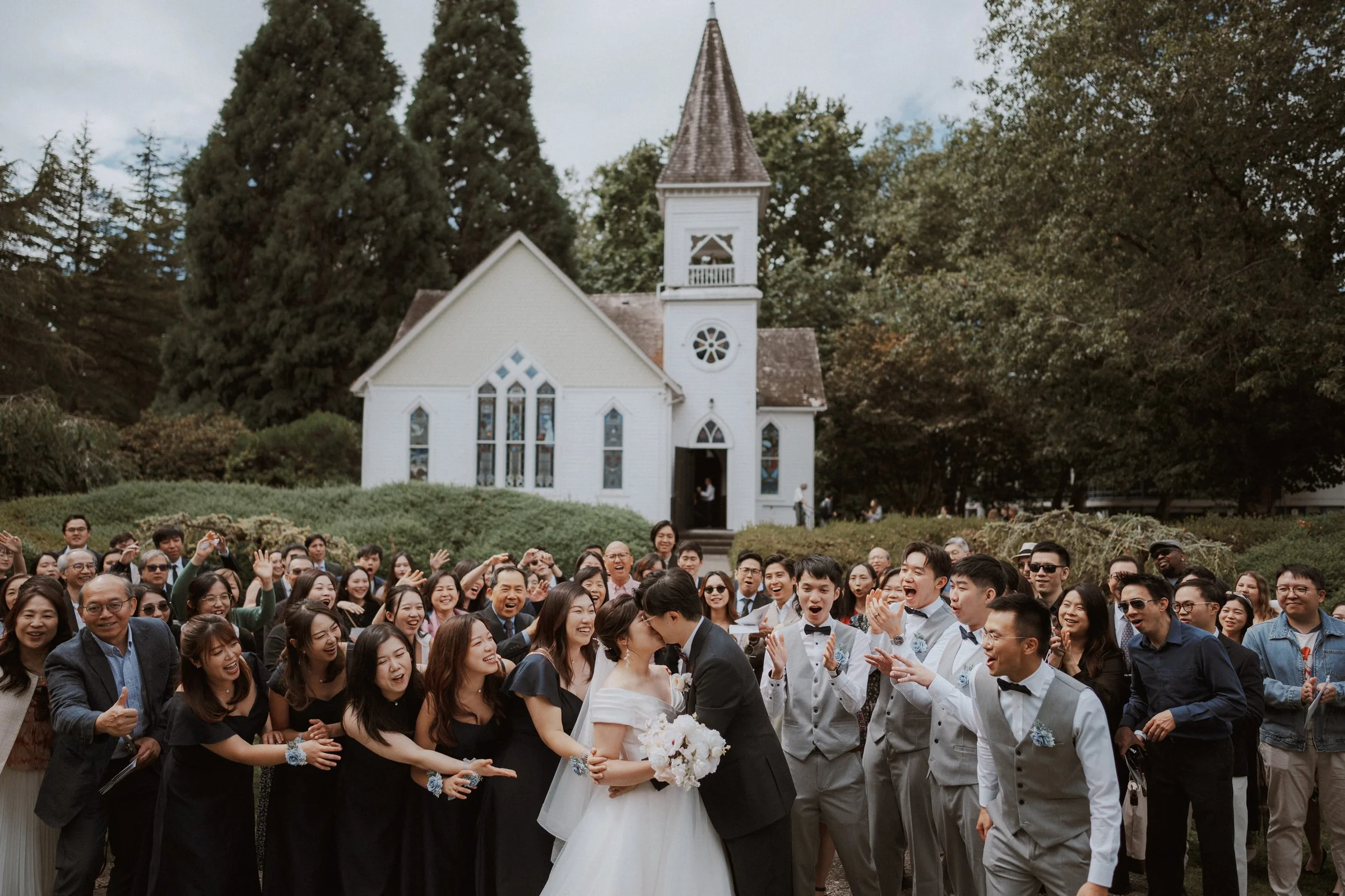 Large wedding group photo outside Minoru Chapel, couple kissing with guests cheering, Richmond BC.