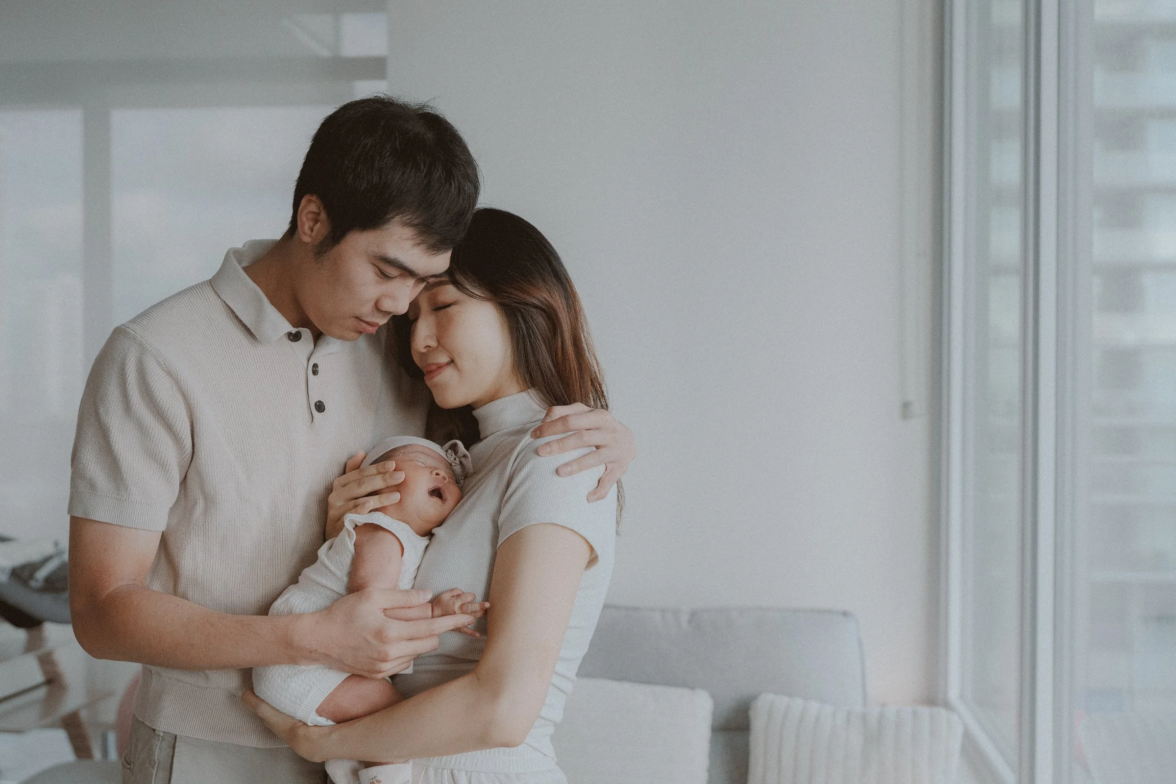 Parents standing together with their newborn beside a softly lit window.