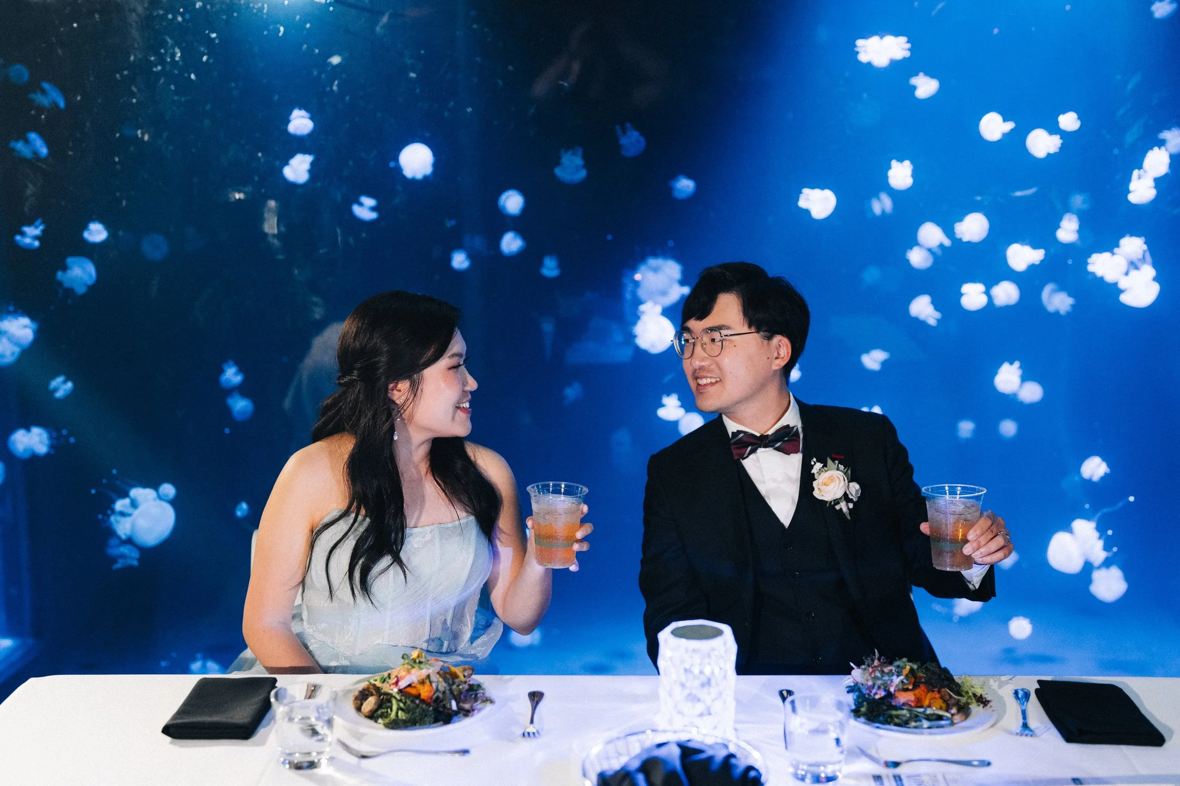 Bride and groom holding drinks at their table in front of a jellyfish display.