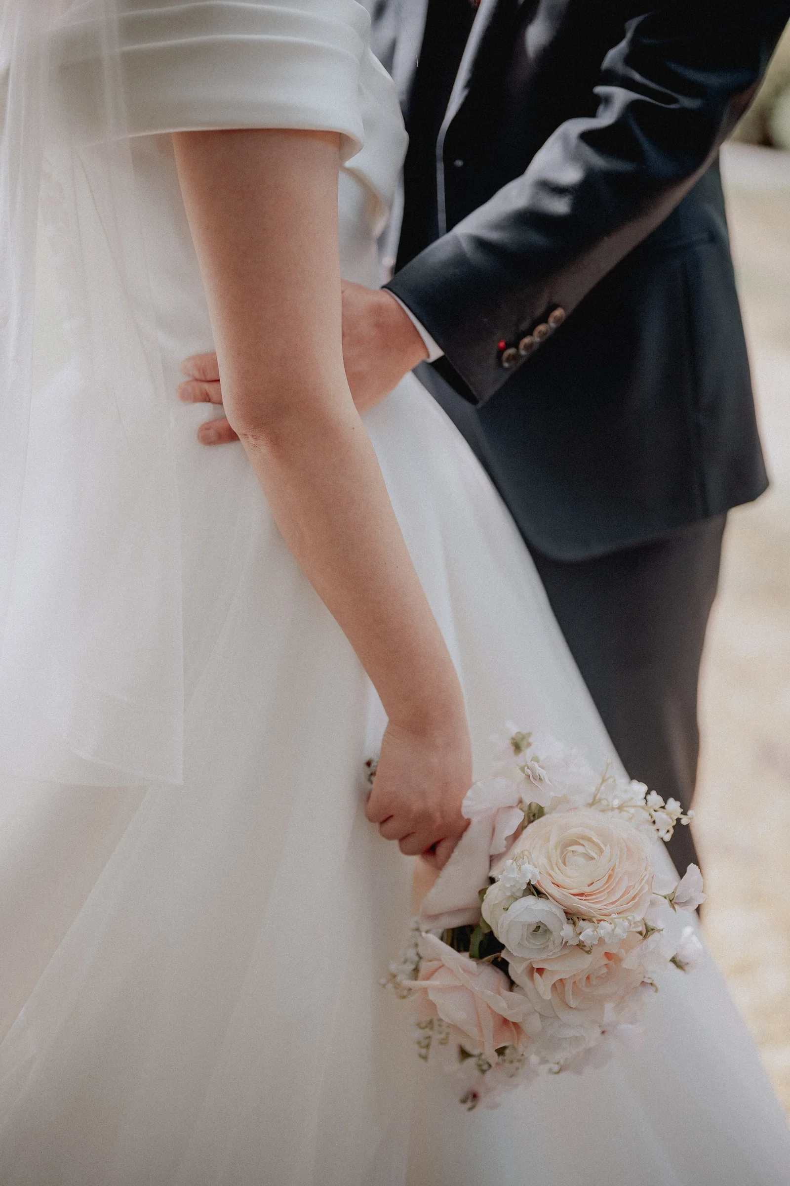 Detail of the bride's bouquet and the couple's hands beside her wedding gown.