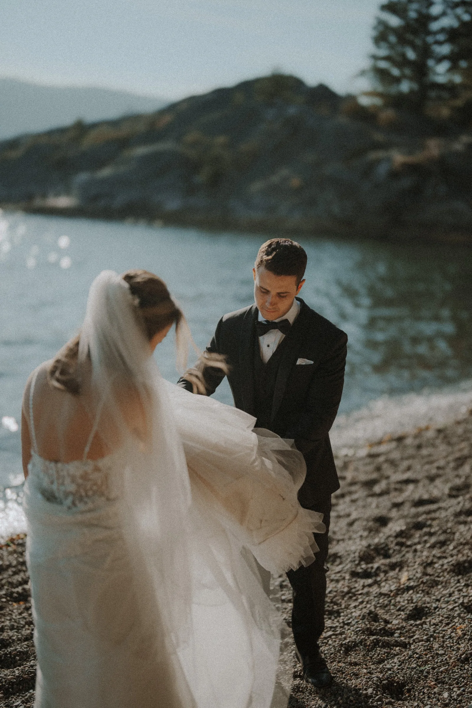 Groom helping hold the bride's gown above the pebbled shoreline.