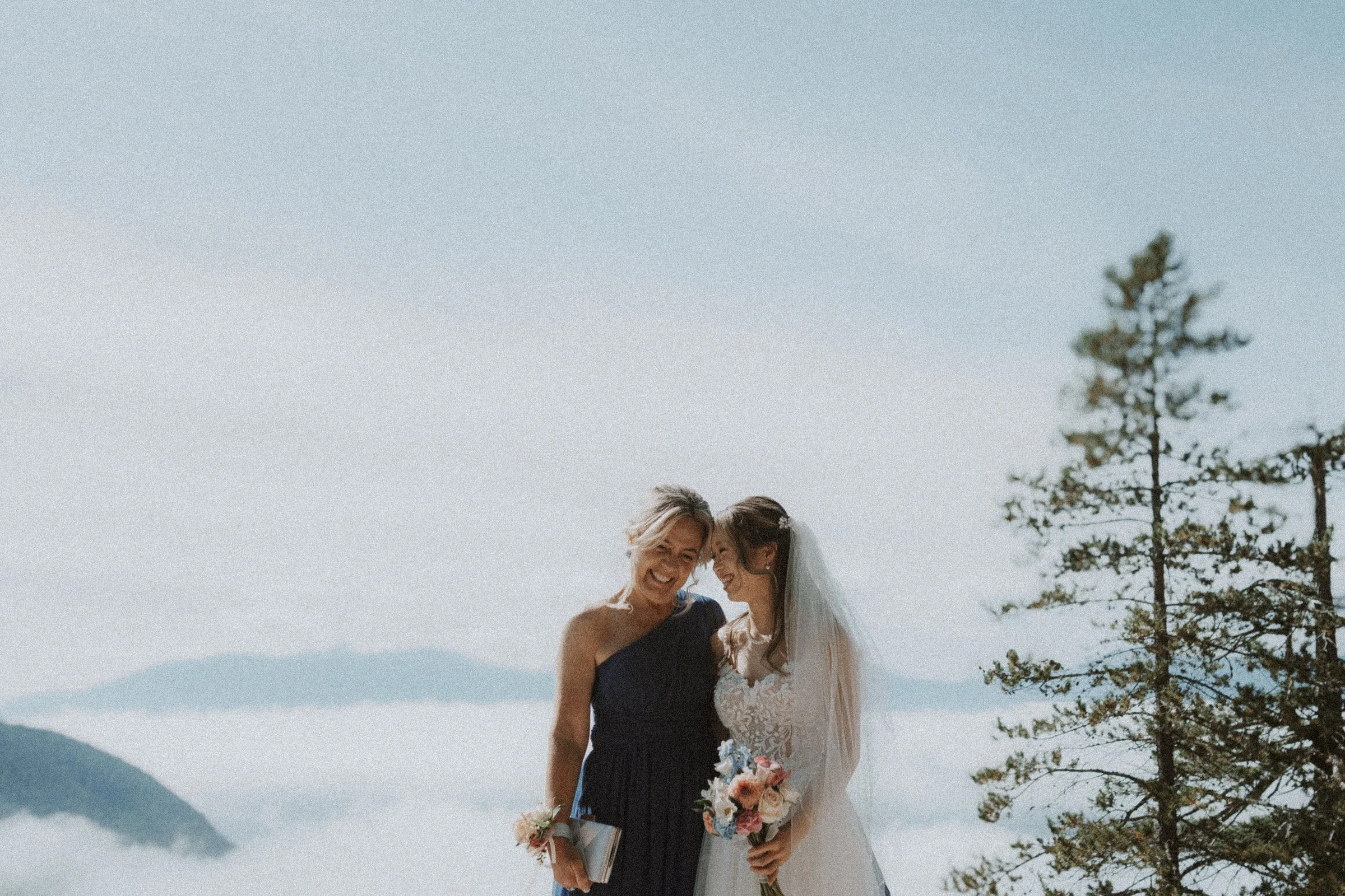 Bride leaning toward a guest as they pose together with the cloud-covered mountains behind.