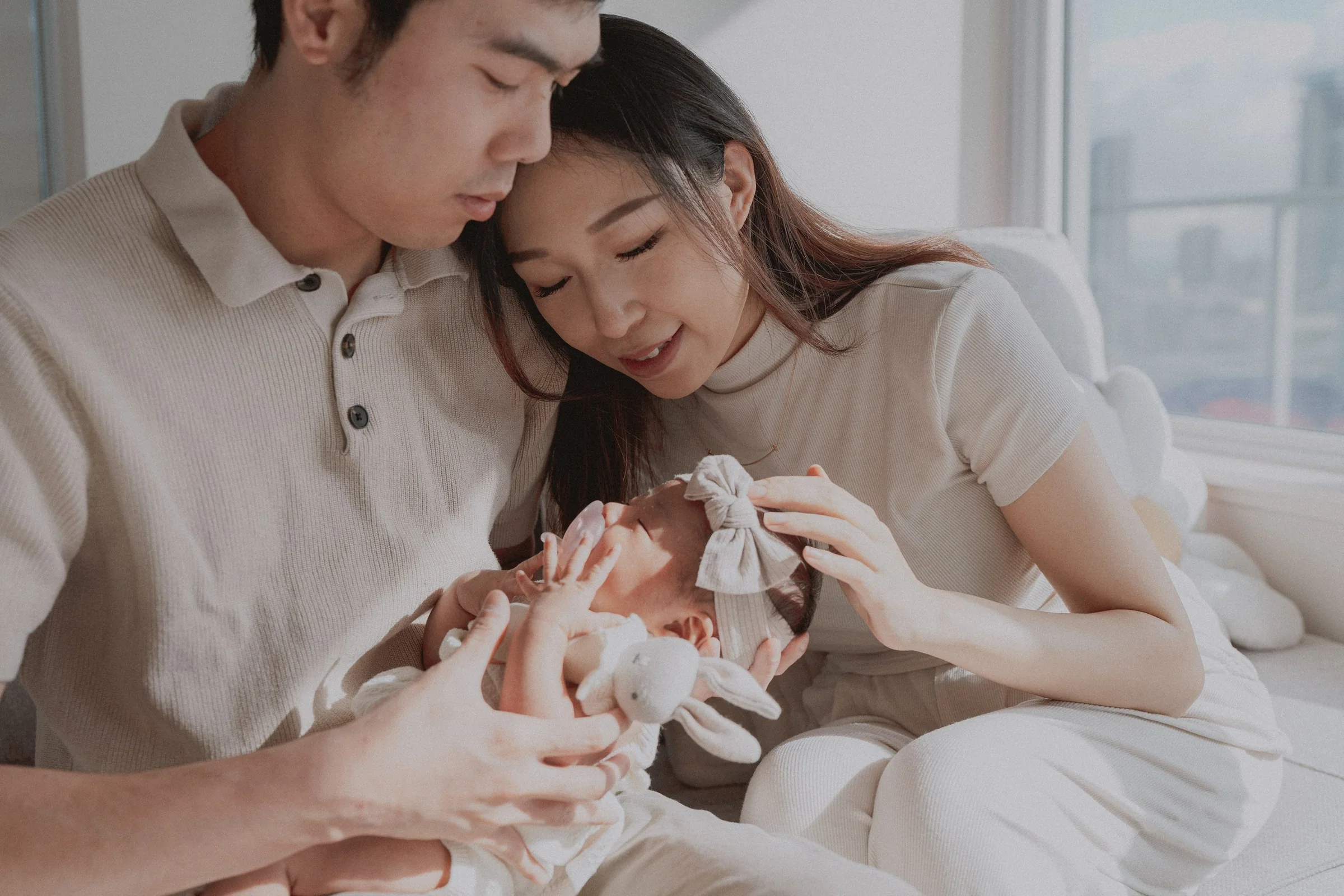 Parents smiling down at their newborn beside a softly lit window.