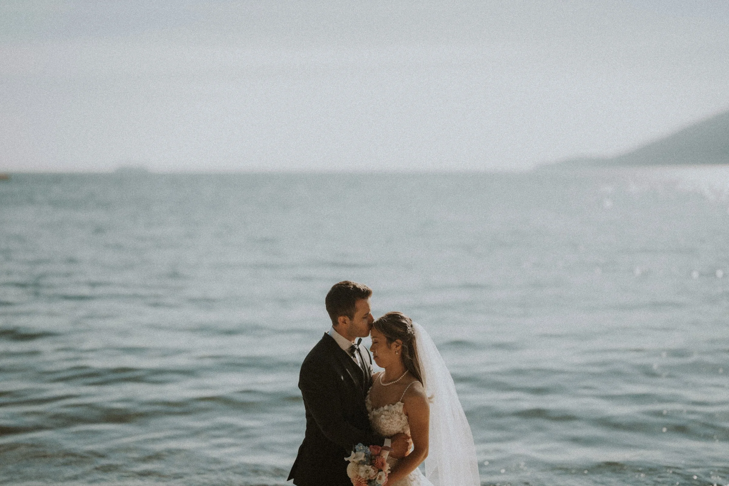 Bride and groom embracing beside calm water at Whytecliff Park.