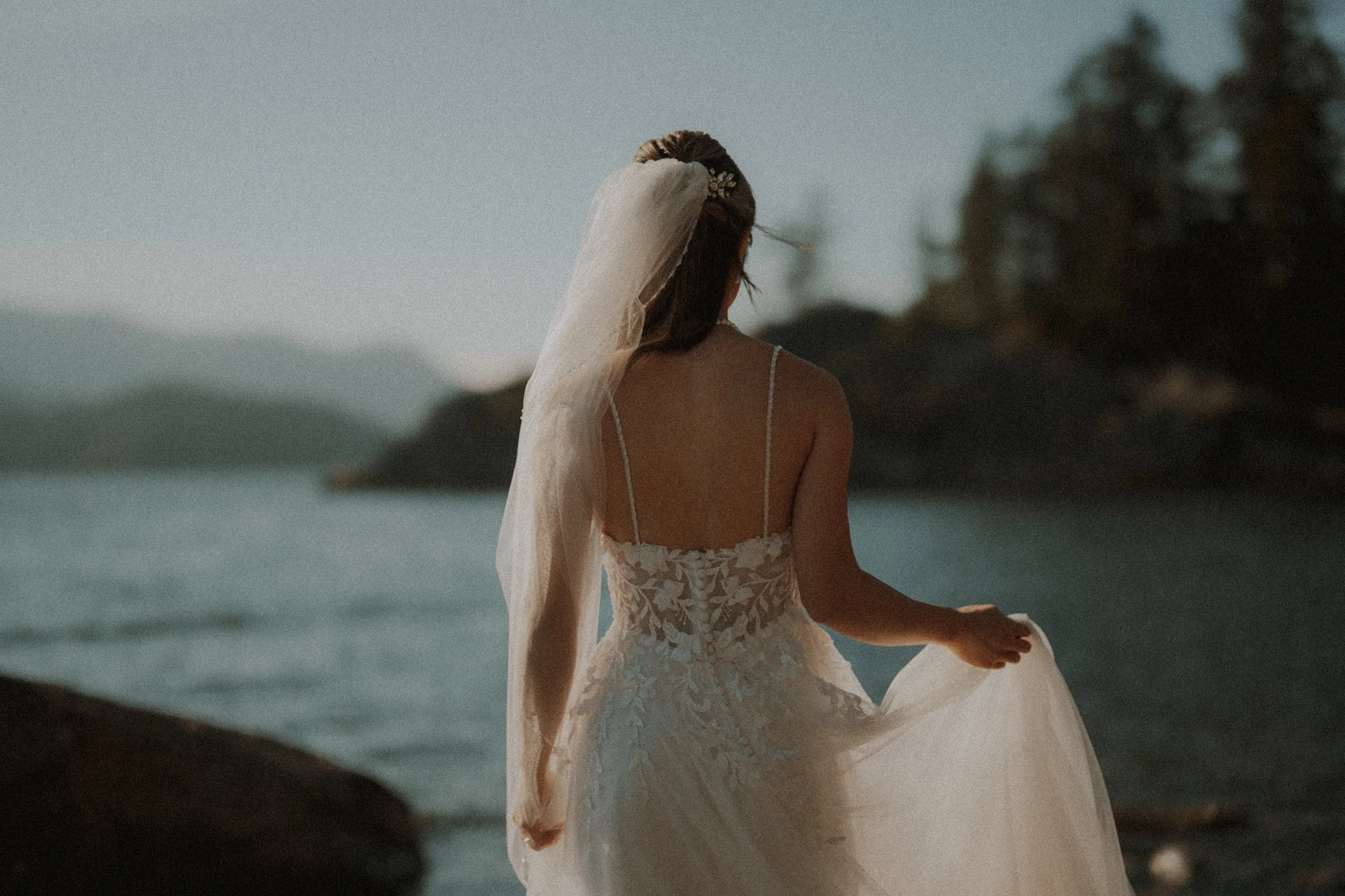 Bride viewed from behind with her veil and gown beside the water.